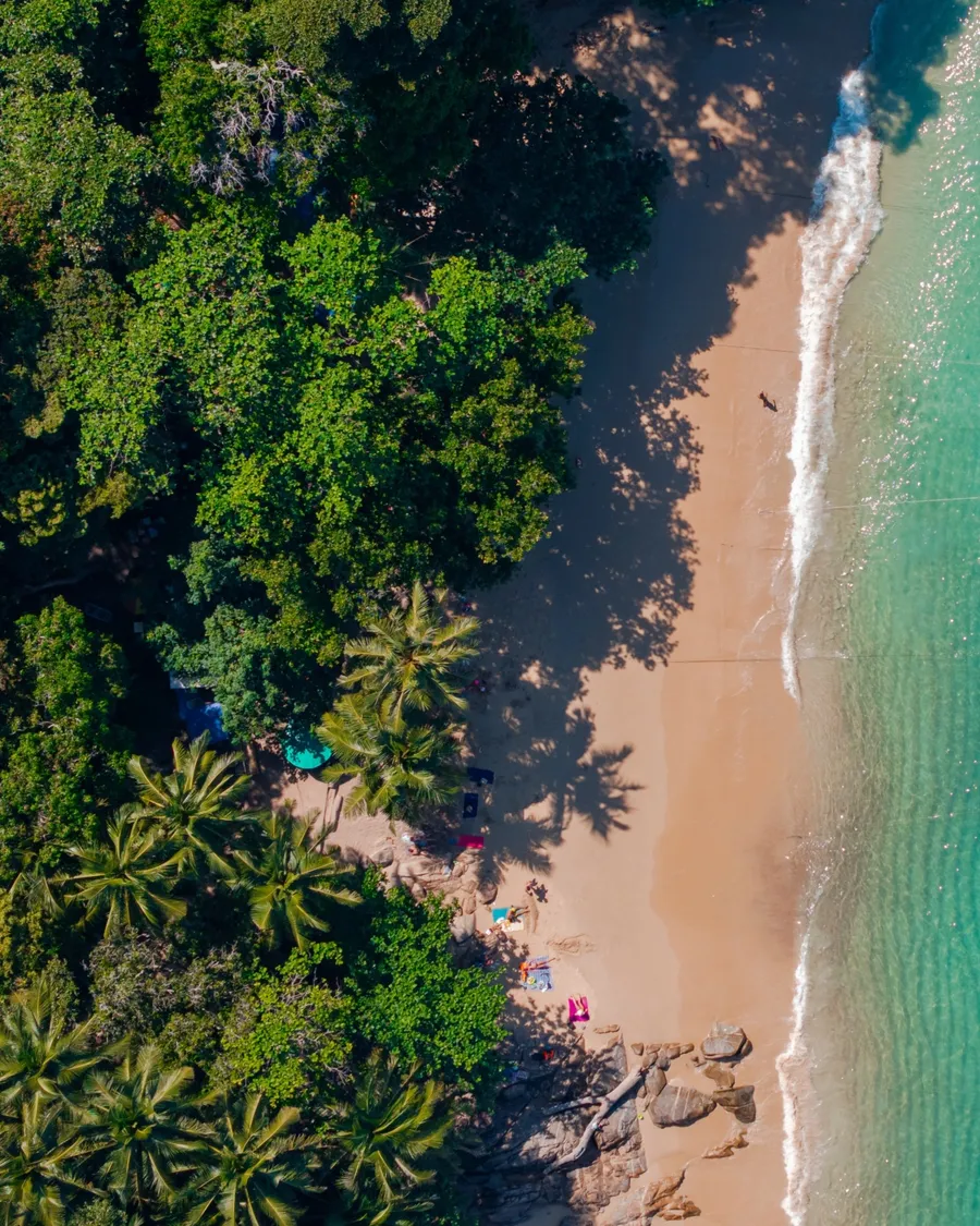 ภาพมุมสูงของอ่าวน้ำใสสีเทอร์คอยซ์ มีเรือหางยาวจอดเรียงกันริมหาดทรายและป่าเขียวทึบด้านซ้าย
